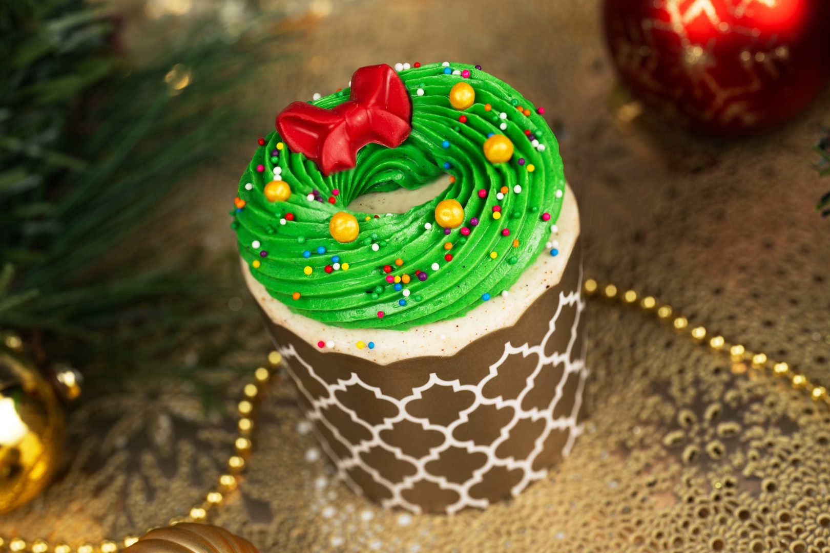 Coquito Wreath Cupcake: Coquito custard-filled vanilla cupcake topped with coquito buttercream, chocolate bow, gold crispy pearls, and rainbow sprinkles  
