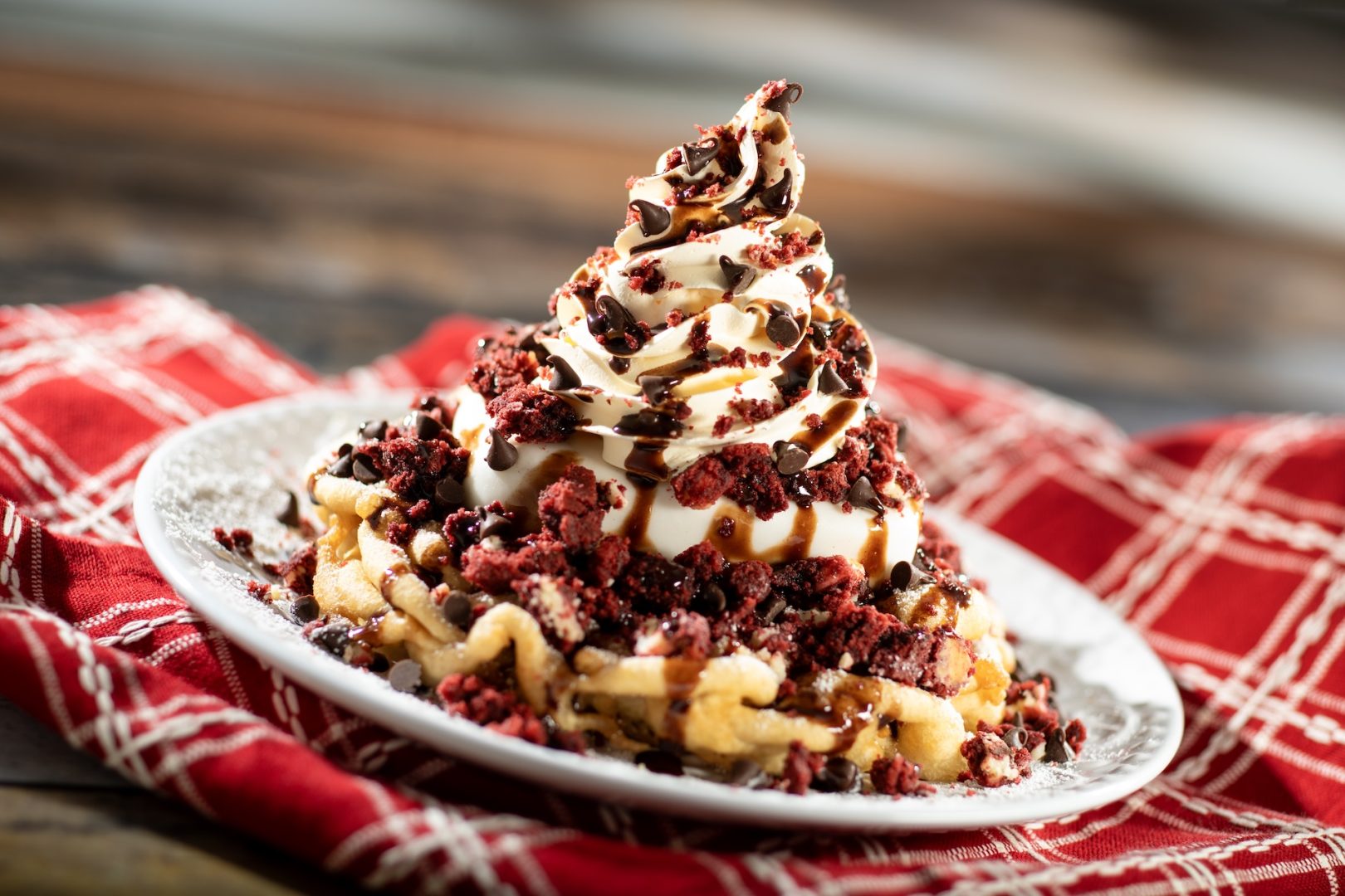 Red Velvet Cookie Funnel Cake topped with powdered sugar, vanilla ice cream, cream cheese whipped cream, red velvet cookie pieces, mini chocolate chips, and chocolate syrup (New)