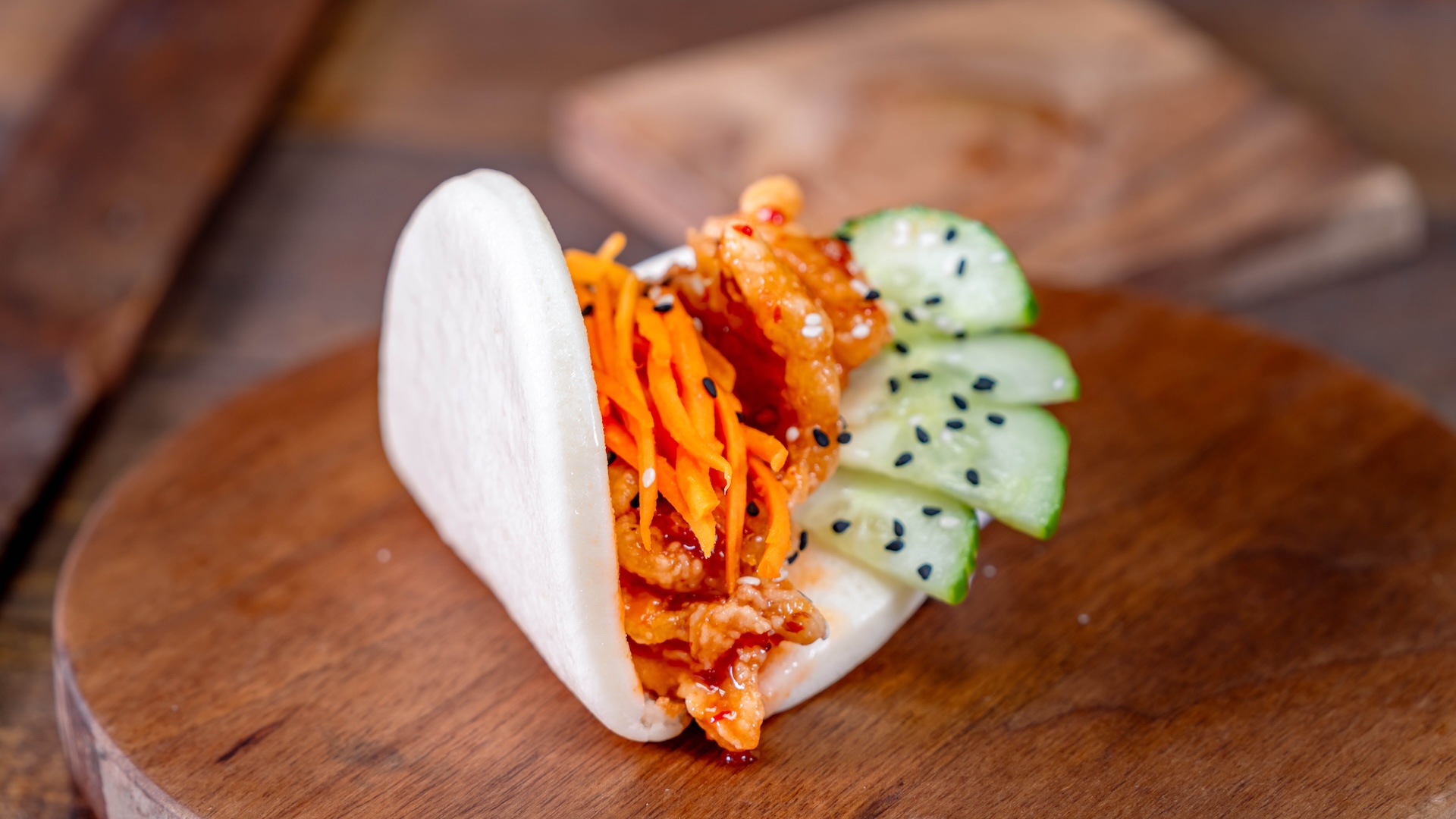 Crispy Chicken Bao: Fried chicken coated in sweet chile sauce with cucumbers, carrots, and sesame seeds served in a soft bao bun (New)