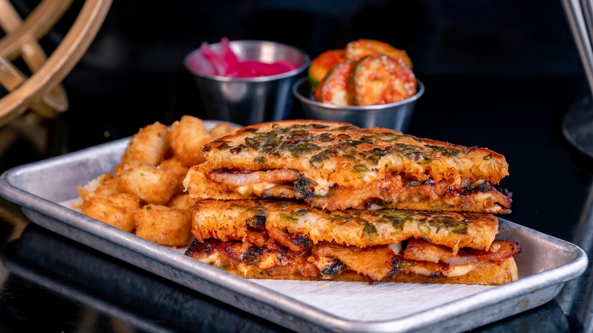 Spicy Pork Belly Grilled Cheese: Pork belly marinated in Korean spices with white American cheese, spicy aïoli, pickled onions, sourdough bread, and green onion crust served with potato bites (New)