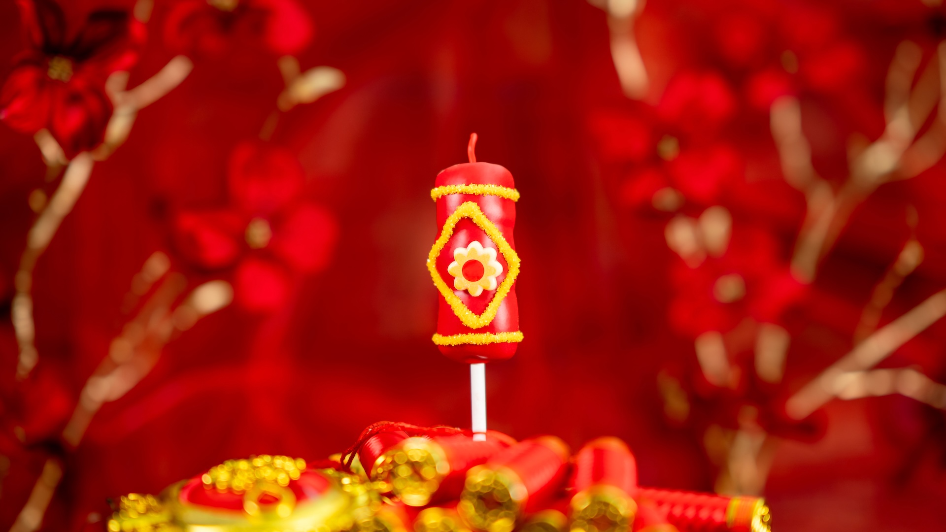 Firecracker Marshmallow Wand: Marshmallow on a stick dipped in caramel and red-colored white chocolate decorated with yellow sanding sugar, a piece of red licorice, yellow-colored white chocolate, and a gold and red flower chocolate piece 